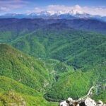 Snow-capped mountains rise above green forested valleys with a winding river road below.