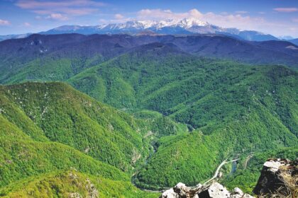 Snow-capped mountains rise above green forested valleys with a winding river road below.