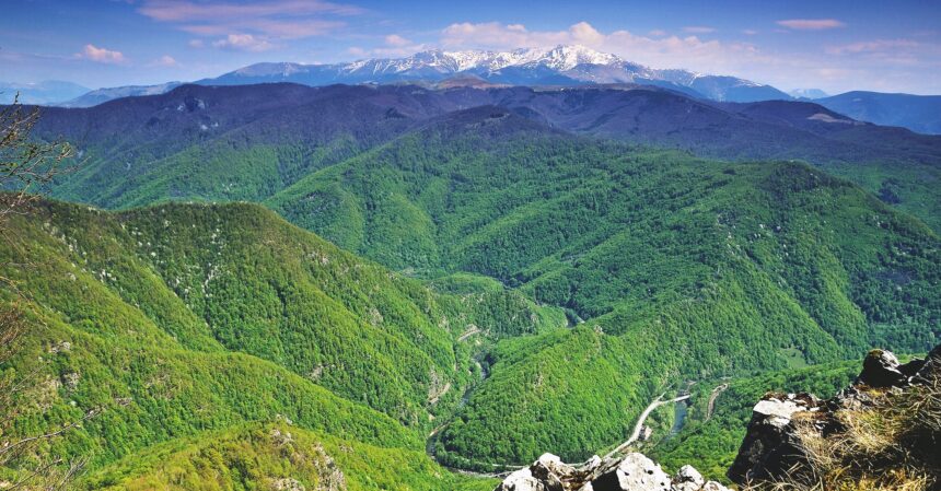 Snow-capped mountains rise above green forested valleys with a winding river road below.