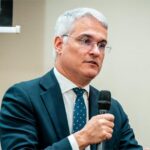 Middle-aged man in a dark teal suit and patterned tie speaks into a handheld microphone at a conference panel.
