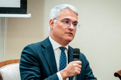 Middle-aged man in a dark teal suit and patterned tie speaks into a handheld microphone at a conference panel.