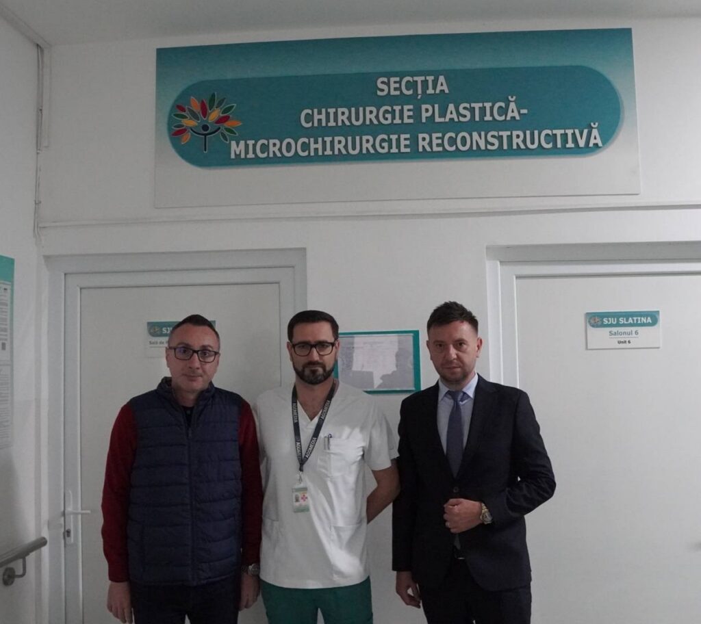 Three men stand in a hospital hallway in front of a sign that reads 'Secția Chirurgie Plastică - Microchirurgie Reconstructivă'.