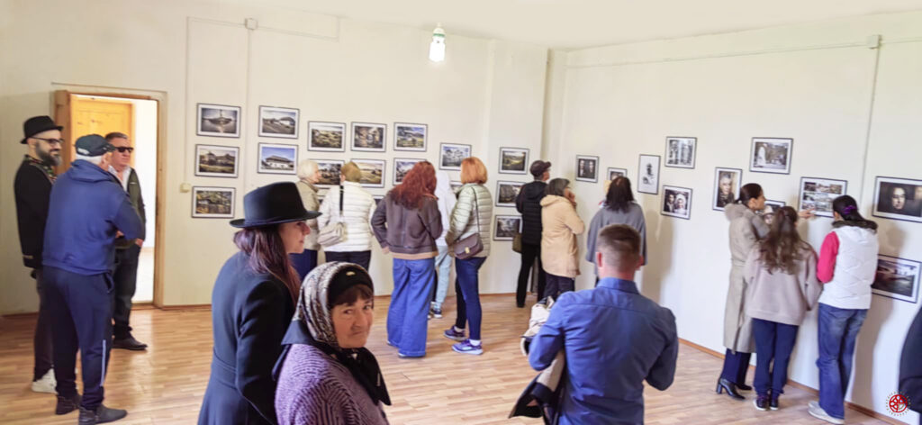 People in a gallery viewing framed photos on white walls, wooden floor, and natural light from above.