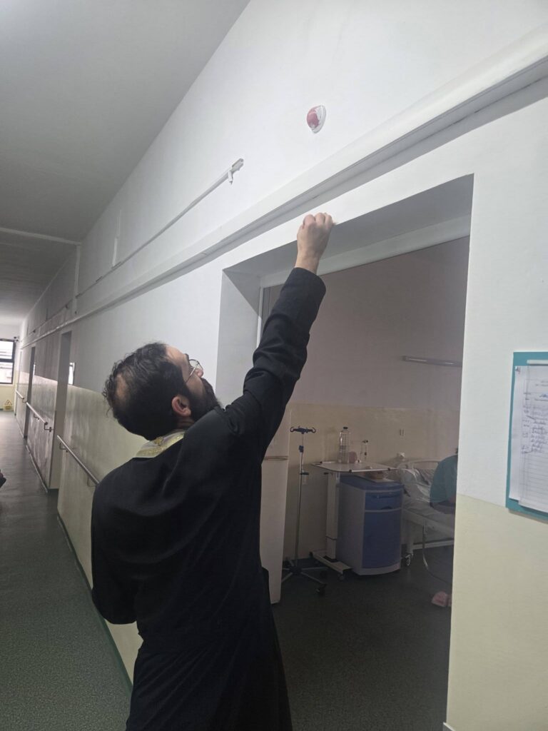 Bearded man in a dark coat stretching his arm up to touch the top of a doorway in a hospital hallway.