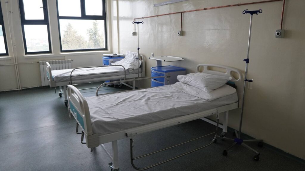 Hospital ward with two empty patient beds, IV stands, and blue bedside cabinets under large windows.