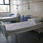 Hospital ward with two empty patient beds, IV stands, and blue bedside cabinets under large windows.
