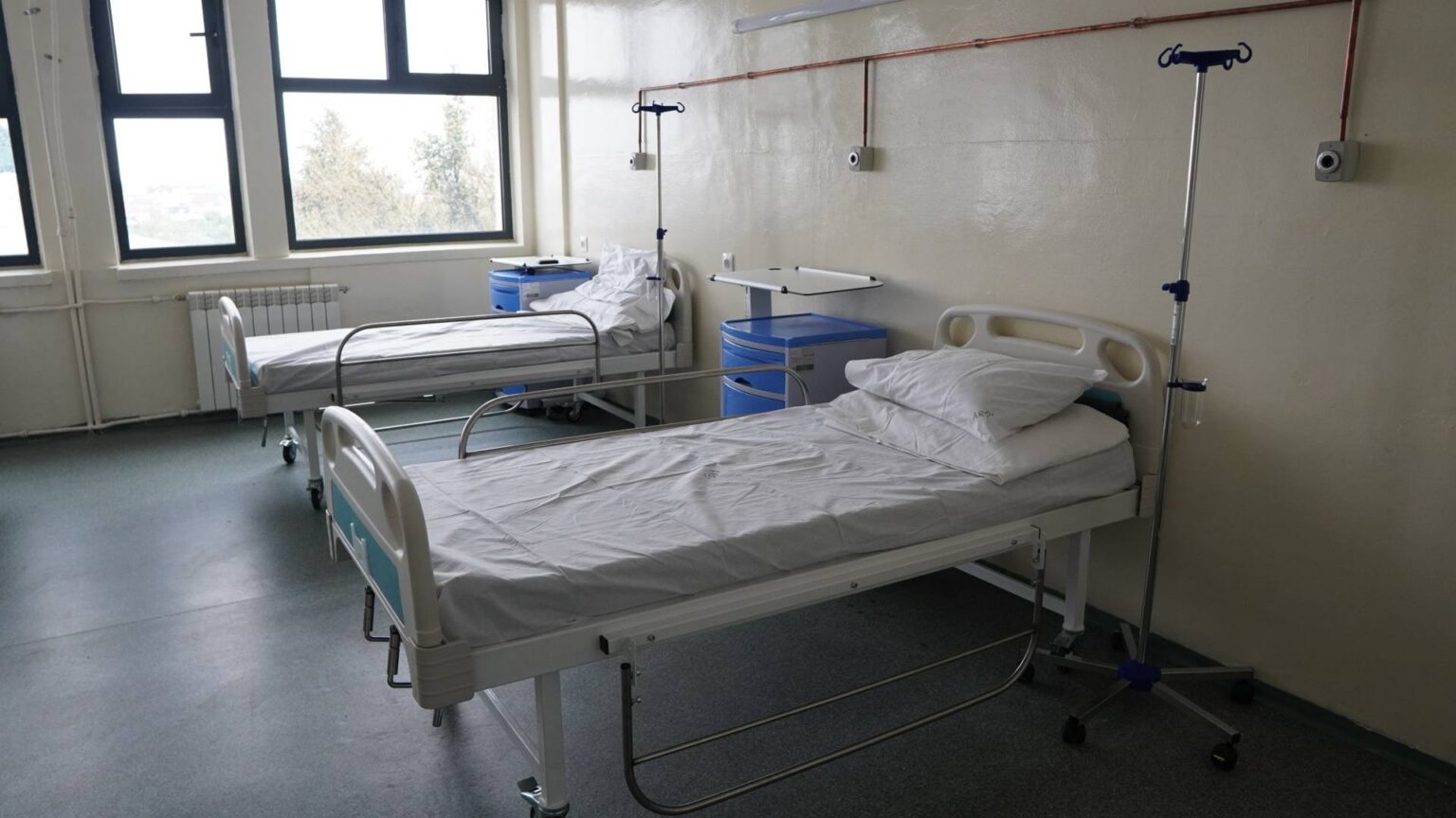 Hospital ward with two empty patient beds, IV stands, and blue bedside cabinets under large windows.