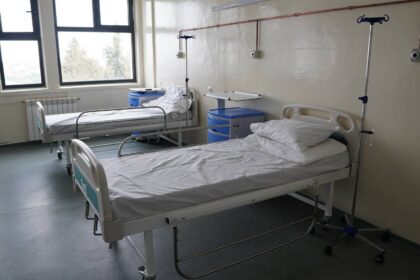 Hospital ward with two empty patient beds, IV stands, and blue bedside cabinets under large windows.