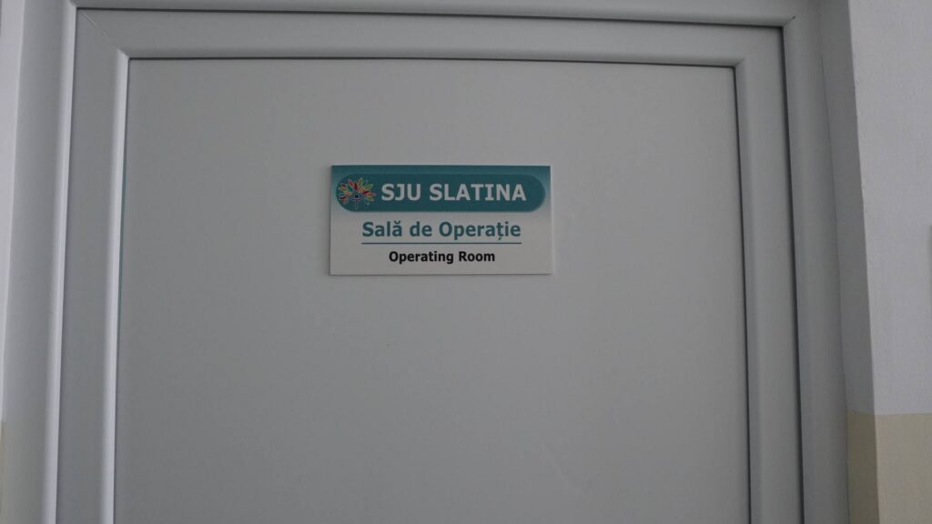 Door sign: SJU Slatina, Sala de Operaţie (Operating Room) with teal banner and flower logo above the doorframe.