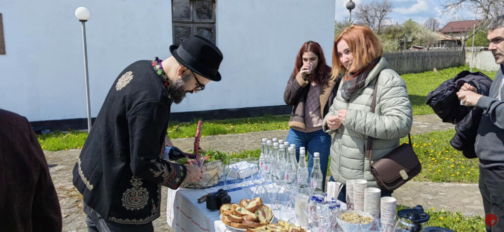 Man in a black hat and embroidered jacket pours a drink at an outdoor table set with bottles, glasses, and snacks while three people watch.