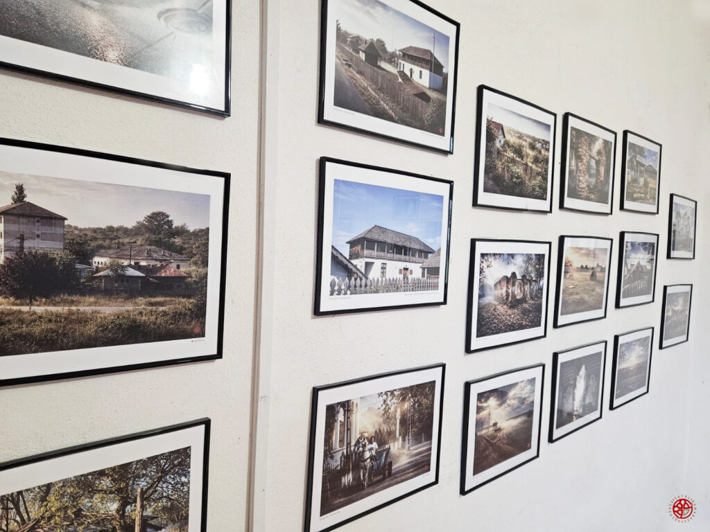 White gallery wall filled with many framed photos arranged in rows, depicting rural houses, landscapes, and trees in an exhibition setting.