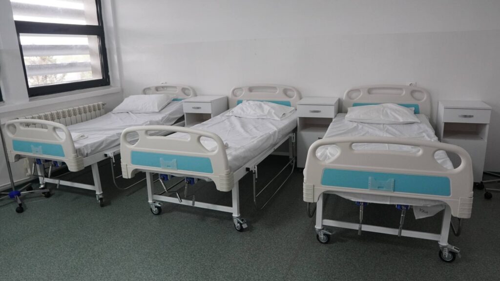 Four hospital beds in a clean, bright ward with white and teal accents and bedside tables between them.