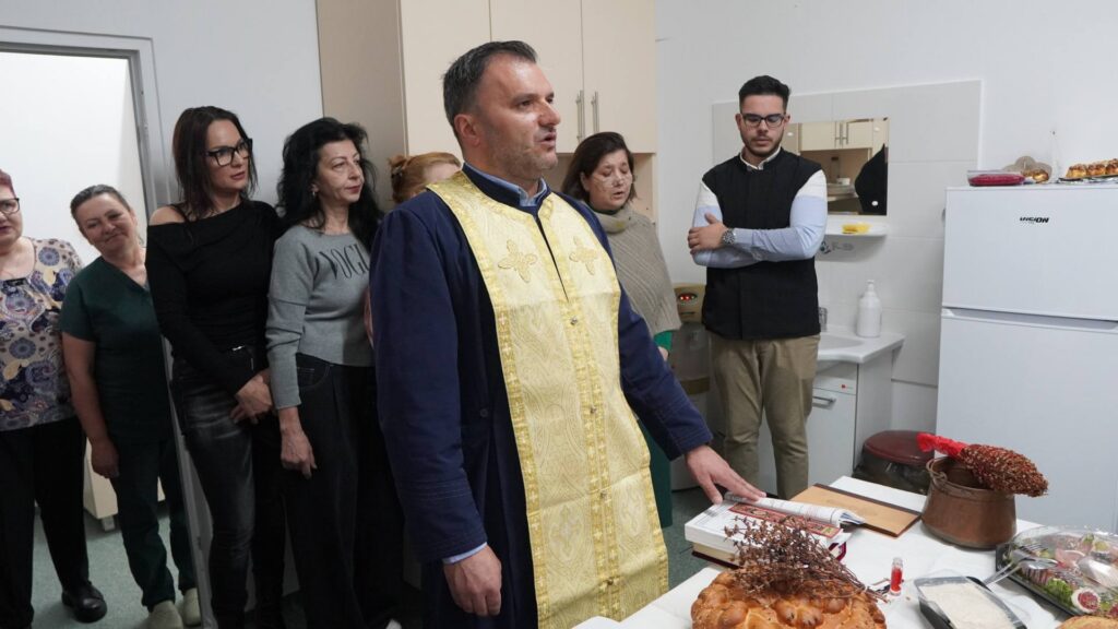 Priest in gold-embroidered vestment leads a ceremony as a line of people stand in a kitchen near a table with bread and pastries behind him.