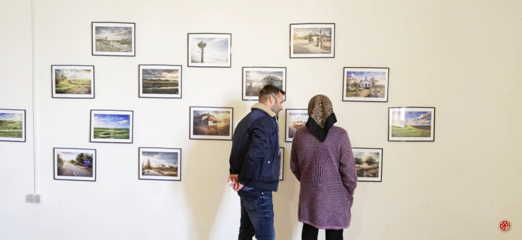 Two people stand side by side, looking at a wall of small framed landscape photographs in a gallery.