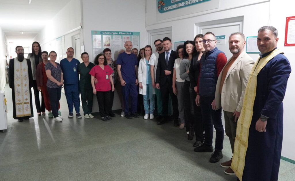 Group of hospital staff, doctors and nurses in scrubs and coats, pose for a group photo in a hospital corridor with two priests in ceremonial robes at each end.