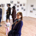 People in a gallery looking at framed photographs hung on white walls; a child in a pink Minnie Mouse hoodie stands near a smiling woman with glasses in the foreground.