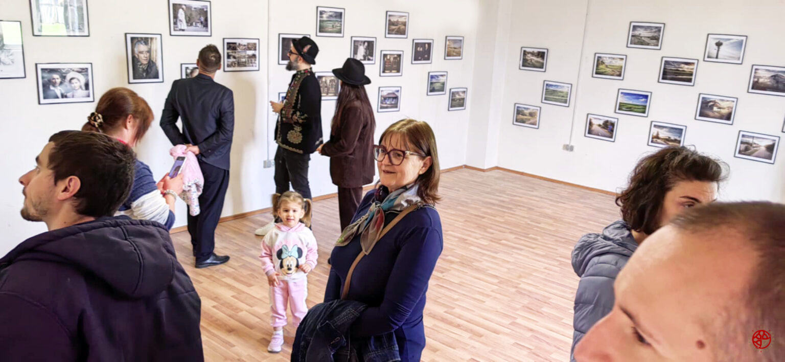 People in a gallery looking at framed photographs hung on white walls; a child in a pink Minnie Mouse hoodie stands near a smiling woman with glasses in the foreground.