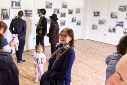 People in a gallery looking at framed photographs hung on white walls; a child in a pink Minnie Mouse hoodie stands near a smiling woman with glasses in the foreground.
