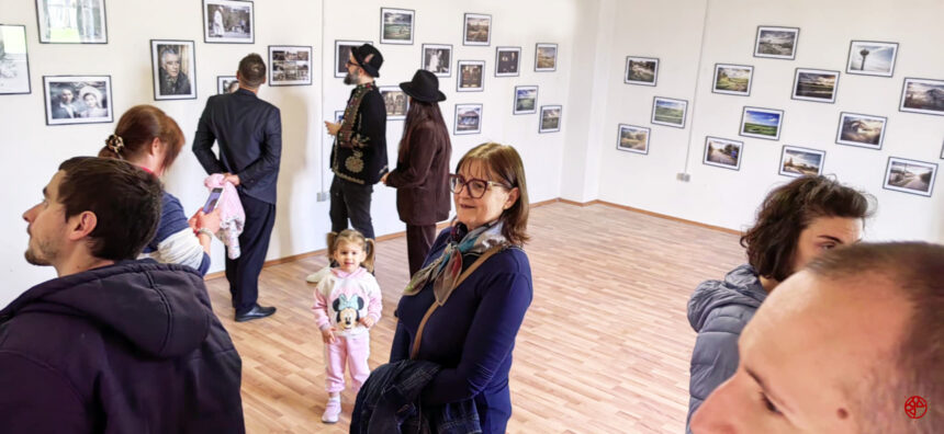 People in a gallery looking at framed photographs hung on white walls; a child in a pink Minnie Mouse hoodie stands near a smiling woman with glasses in the foreground.