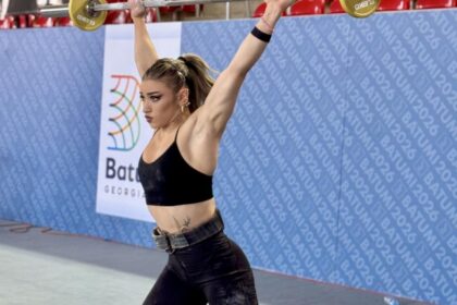 Female weightlifter lifting a barbell overhead during a competition, wearing a black sports bra and dark pants, with blue banners and red seats in the background.