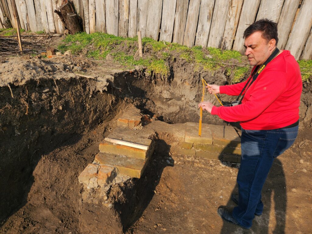Man in a red sweater standing beside a brick-lined excavation trench, measuring with a yellow tape measure.