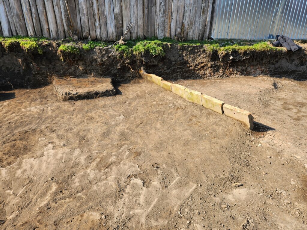 Excavated yard with bare soil, a short concrete step, and a row of rectangular concrete blocks forming a boundary near a wooden fence.