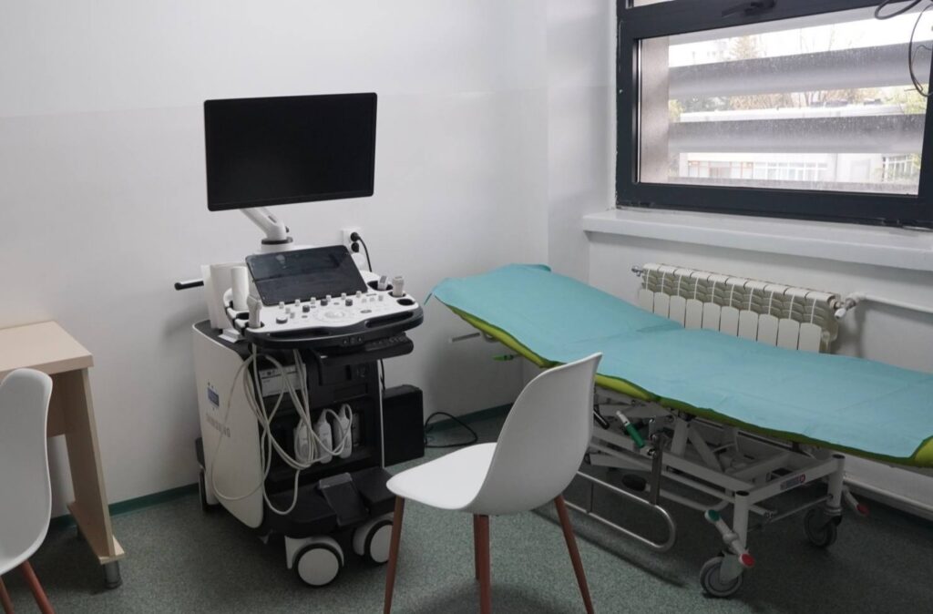 Ultrasound machine with a large monitor and control panel beside an examination bed in a clinical room with a teal sheet.