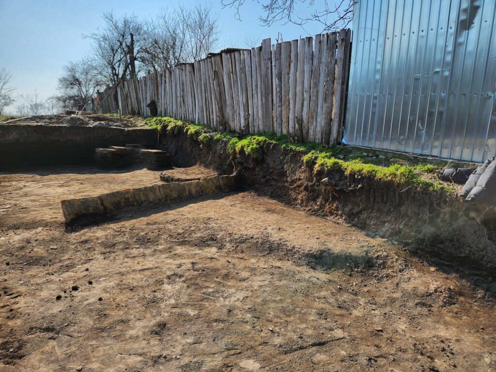 Construction site with a trench along a weathered wooden fence and a metal building on the right, moss growing at the edge of the soil.