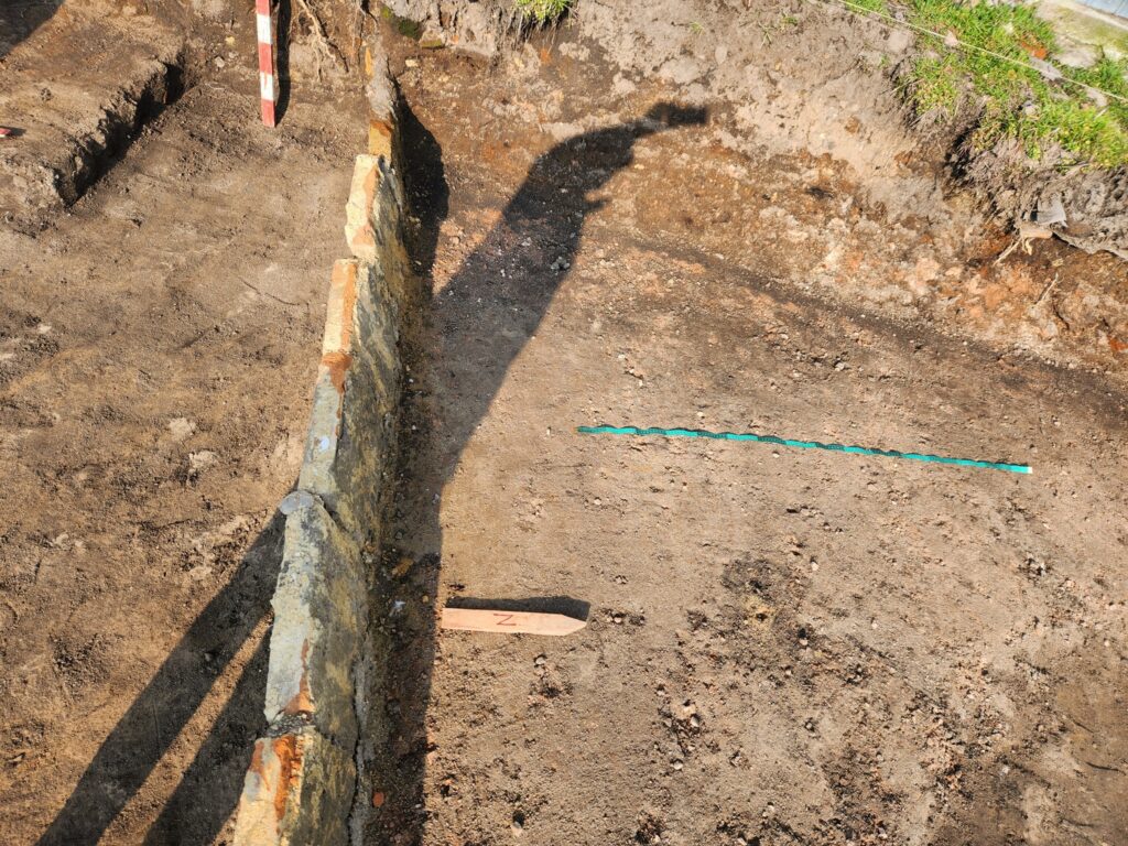 Foundation trench with a stone wall along the left and bare, dusty soil; a teal measuring tape lies across the ground.