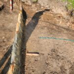 Foundation trench with a stone wall along the left and bare, dusty soil; a teal measuring tape lies across the ground.