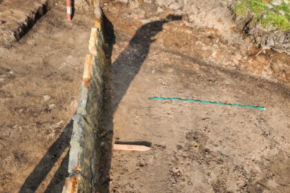 Foundation trench with a stone wall along the left and bare, dusty soil; a teal measuring tape lies across the ground.