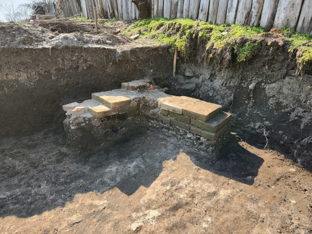 Excavation pit with a small brick platform stacked inside the dirt, near a weathered wooden fence edged with mossy soil.