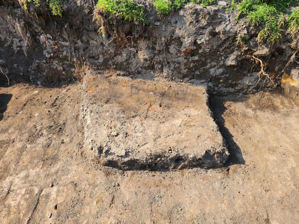 Rectangular concrete block partially buried in compacted dirt at a construction site, with a soil wall and exposed roots above.