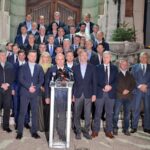 A man stands at a glass podium with microphones, flanked by a large group of formally dressed people on stone stairs outside a building, as if at a press conference.
