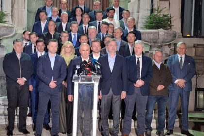 A man stands at a glass podium with microphones, flanked by a large group of formally dressed people on stone stairs outside a building, as if at a press conference.