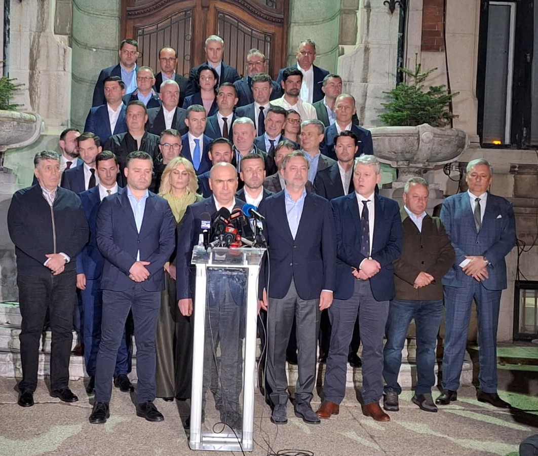 A man stands at a glass podium with microphones, flanked by a large group of formally dressed people on stone stairs outside a building, as if at a press conference.