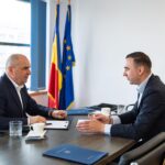 Two men in business suits sit at a conference table, discussing documents with blue folders and coffee cups, flags in the background.