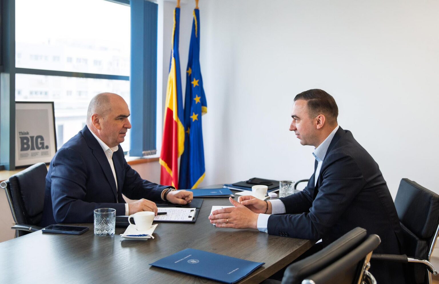 Two men in business suits sit at a conference table, discussing documents with blue folders and coffee cups, flags in the background.