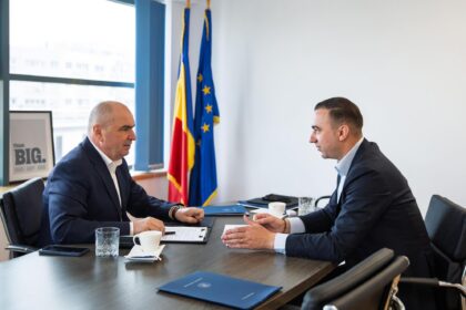 Two men in business suits sit at a conference table, discussing documents with blue folders and coffee cups, flags in the background.