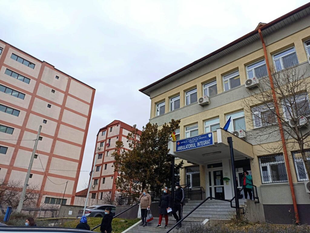 People wearing face masks walk down the stairs outside a hospital building with beige walls, red trim, and a blue sign.