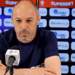 Man at a press conference wearing a navy shirt, speaking into a microphone with sponsor banners in the background and two water bottles on the table.