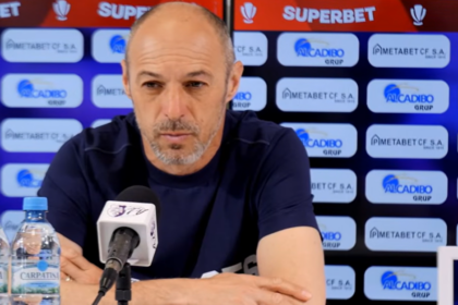Man at a press conference wearing a navy shirt, speaking into a microphone with sponsor banners in the background and two water bottles on the table.