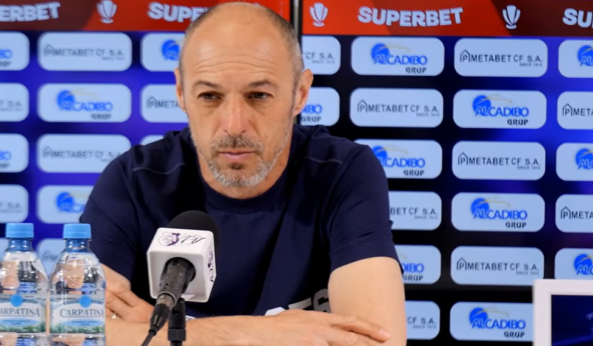 Man at a press conference wearing a navy shirt, speaking into a microphone with sponsor banners in the background and two water bottles on the table.