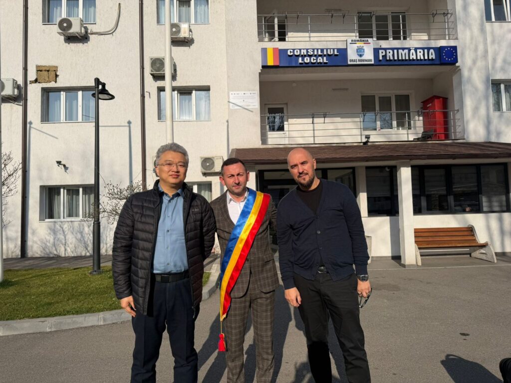 Three men stand outside a local government building; the man in the center wears a Romanian tricolor sash, suggesting an official role.