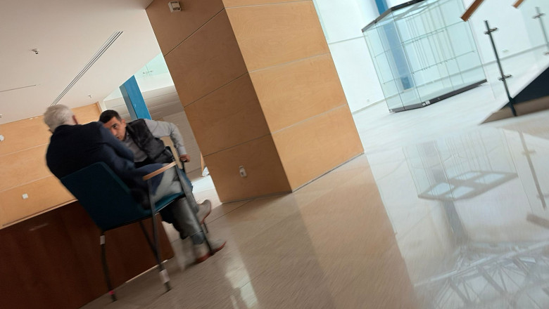 Two men sit facing each other on blue chairs in a modern, wood-paneled lobby with a glossy tiled floor.