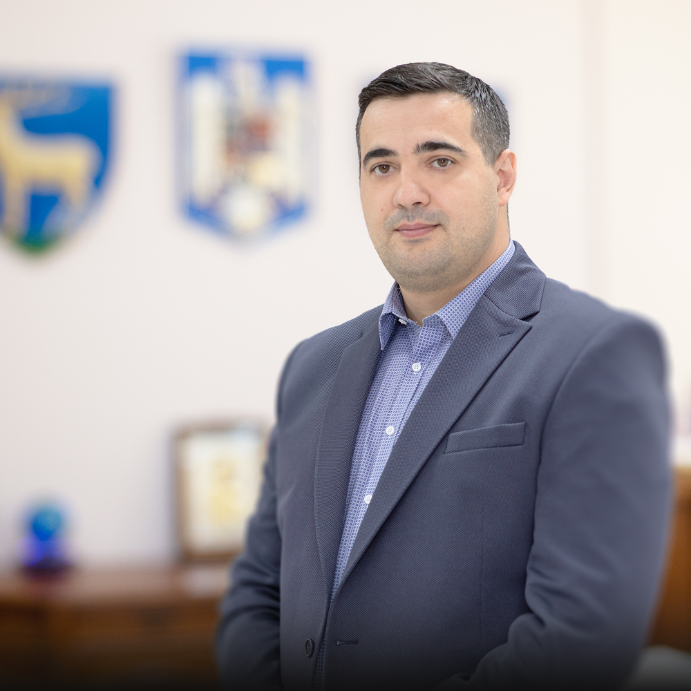 Professional portrait of a man in a navy blazer and blue dress shirt in an office setting with blurred coats of arms in the background
