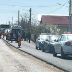 Police and road workers conducting a roadside stop on a rural street, with a police car, a worker in a reflective vest, and traffic cones along the road.