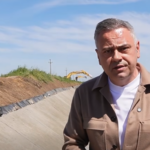 Man in a tan jacket speaks at a construction site, standing in a dug trench with equipment in the background under a blue sky.