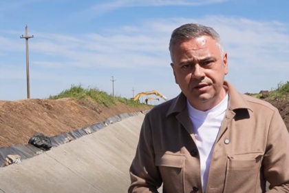 Man in a tan jacket speaks at a construction site, standing in a dug trench with equipment in the background under a blue sky.