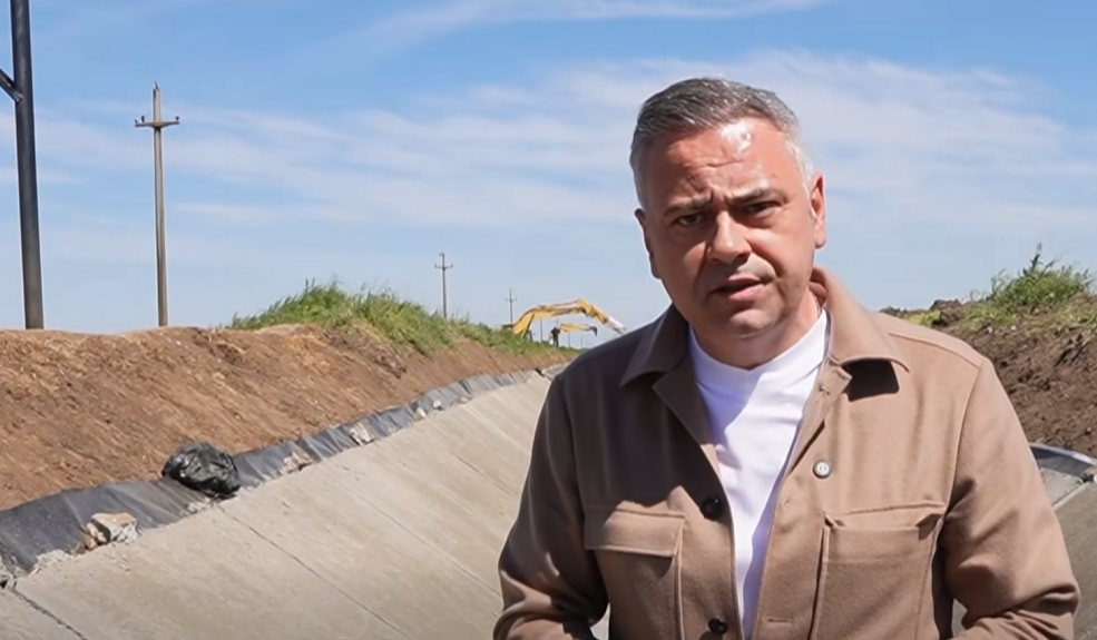 Man in a tan jacket speaks at a construction site, standing in a dug trench with equipment in the background under a blue sky.